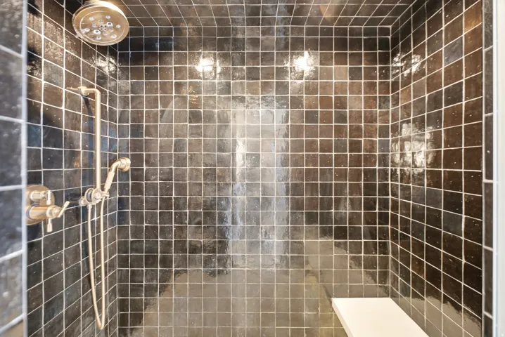 Shower stall featuring dark square tiles, a fixed showerhead, a handheld shower fixture, and a built-in bench