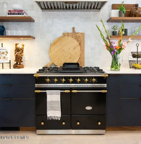 Gourmet kitchen featuring a professional-grade black range with brass accents, light-colored tile backsplash, custom blue cabinetry with dark hardware, white countertops, and open wood shelving