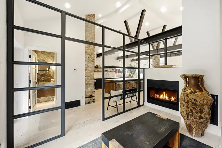 Open-concept living space featuring a modern fireplace, vaulted ceilings with exposed beams, wood-finish flooring, and industrial-style glass room dividers