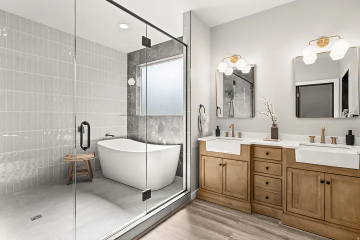 Glass-enclosed shower with vertical subway tile surround, freestanding soaking tub, dual vanity with undermount farmhouse sinks, and light wood-finish flooring