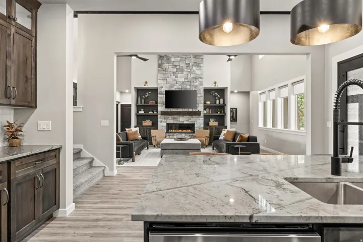 Spacious open-concept interior featuring light-toned flooring, a stone fireplace, built-in shelving, high ceilings, and a kitchen island with an undermount sink and a matte black faucet