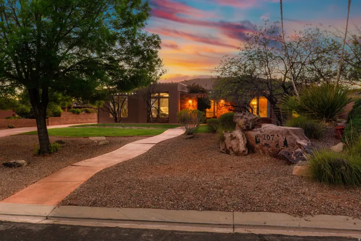 Desert modern architecture featuring an earth-toned exterior, multiple large window arrays, mature landscaping, a curved concrete pathway, and a gravel front yard