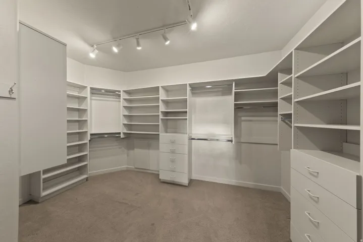 Spacious walk-in closet featuring extensive built-in shelving, multiple hanging rods, integrated drawer units, track lighting, and neutral carpeting
