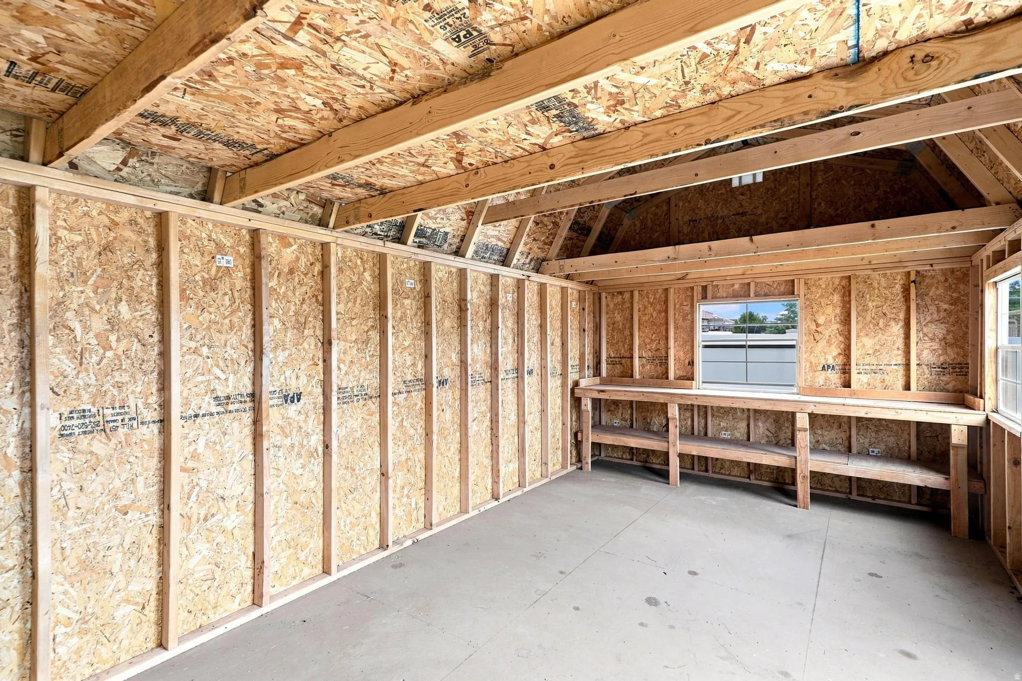 Unfinished interior space featuring exposed wood framing and oriented strand board (OSB) walls and ceiling, a built-in wood workbench with lower shelving, and a concrete floor