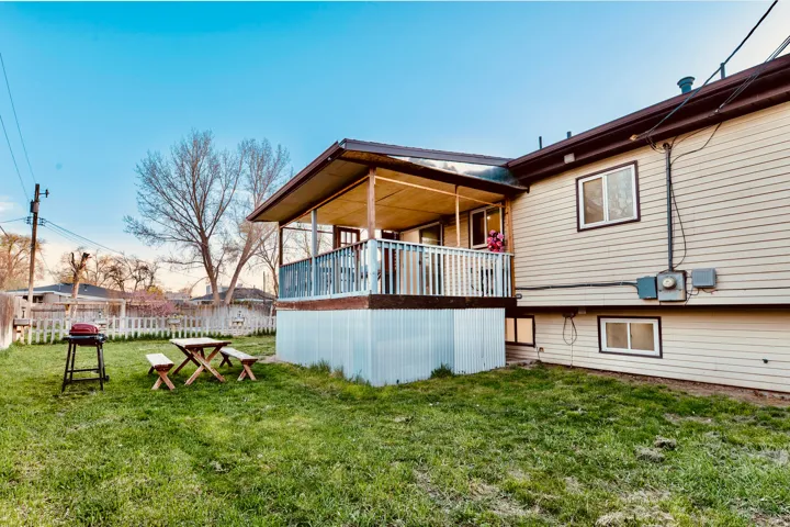 Expansive grassy yard featuring a covered wooden deck with white railings, tan horizontal siding, and multiple windows