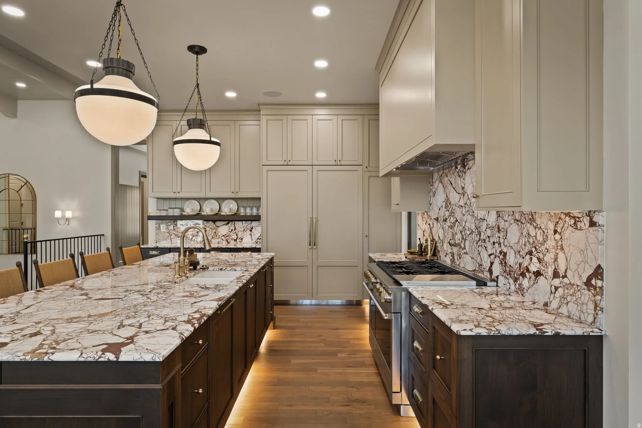 Gourmet kitchen featuring a substantial island with integrated sink, richly veined stone countertops and backsplash, custom cabinetry, stainless steel appliances, and warm wood flooring