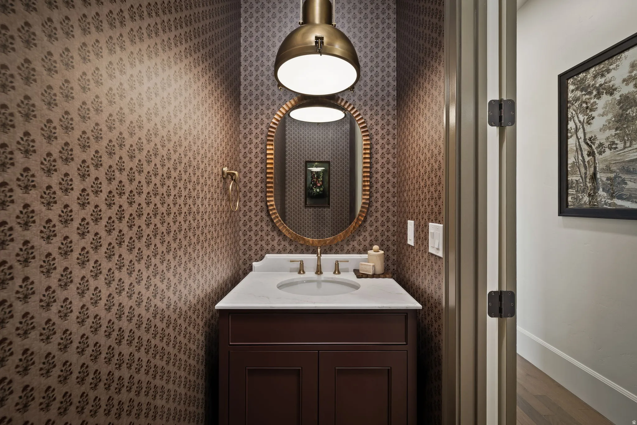 Powder room featuring patterned wallpaper, a dark wood vanity with a white countertop, an oval framed mirror, and a brass pendant light fixture