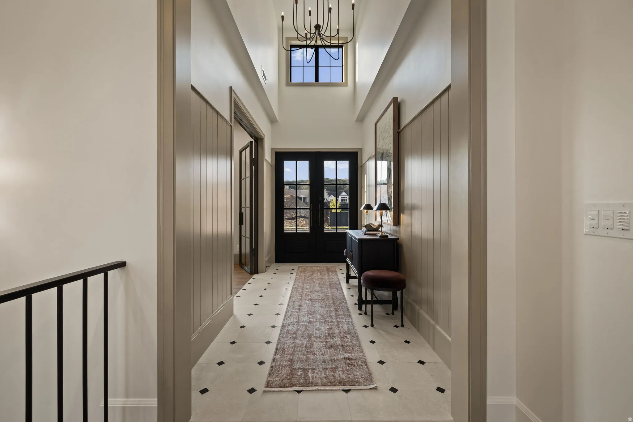 Two-story foyer featuring a black wrought iron chandelier, paneled wainscoting, and a light-toned tile floor with dark accent squares