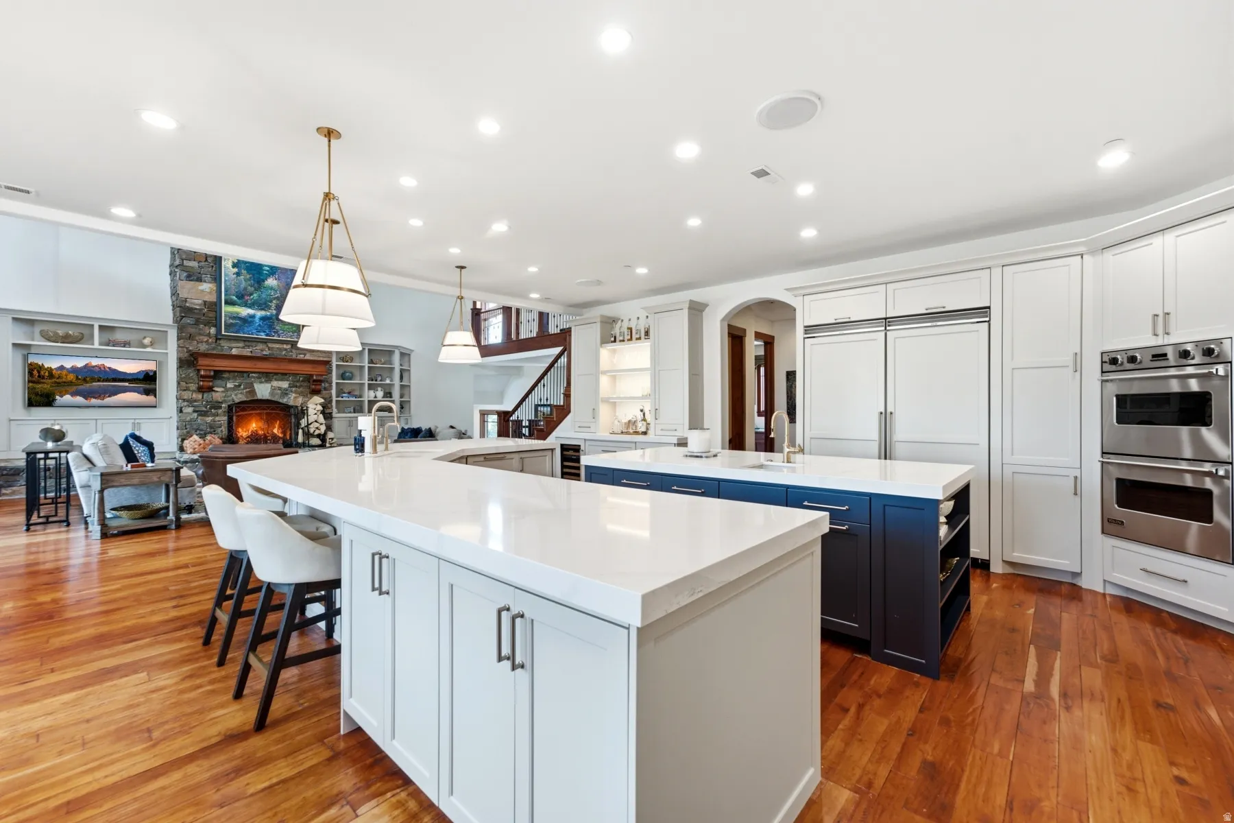 Gourmet kitchen and living area featuring wood-finish flooring, two large kitchen islands with white countertops, integrated paneled refrigerators, stainless steel double ovens, and a stone fireplace with a wood mantel