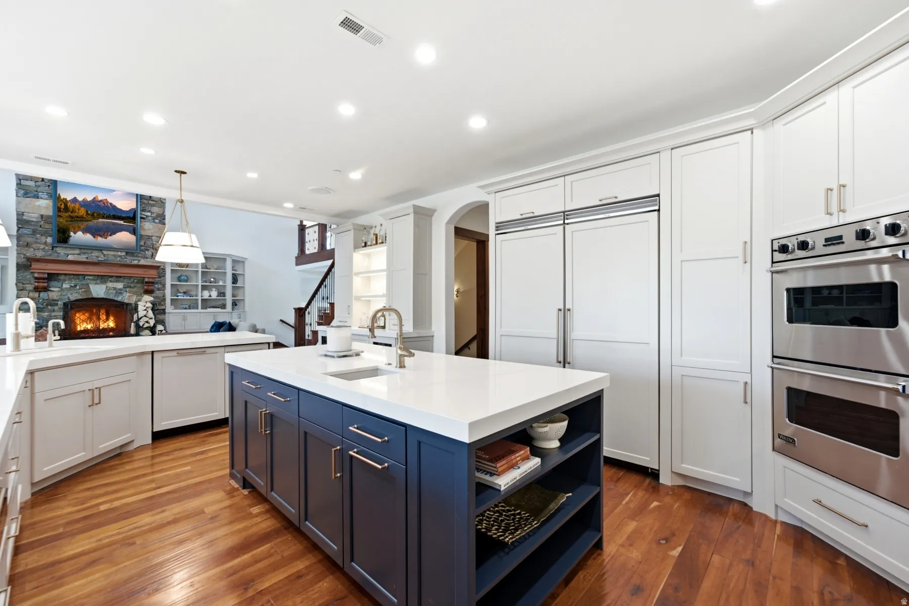 Spacious kitchen featuring a large central island with a white countertop and blue cabinetry, rich wood-finish flooring, integrated paneled refrigerator, stainless steel double ovens, and recessed lighting