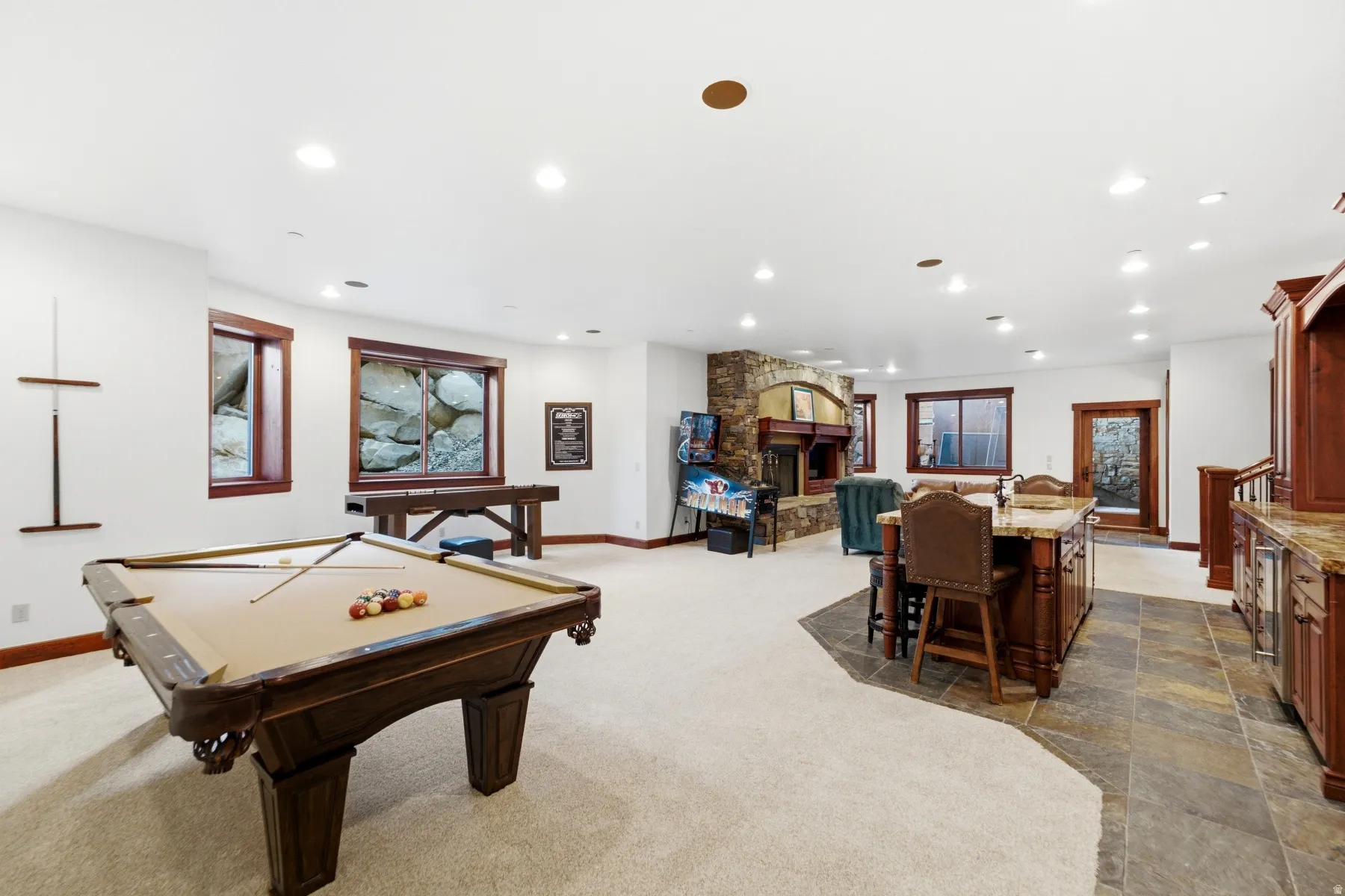 Expansive entertainment area featuring a billiard table, stone-clad fireplace, integrated bar with stone countertops, recessed lighting, and rich wood-trimmed windows