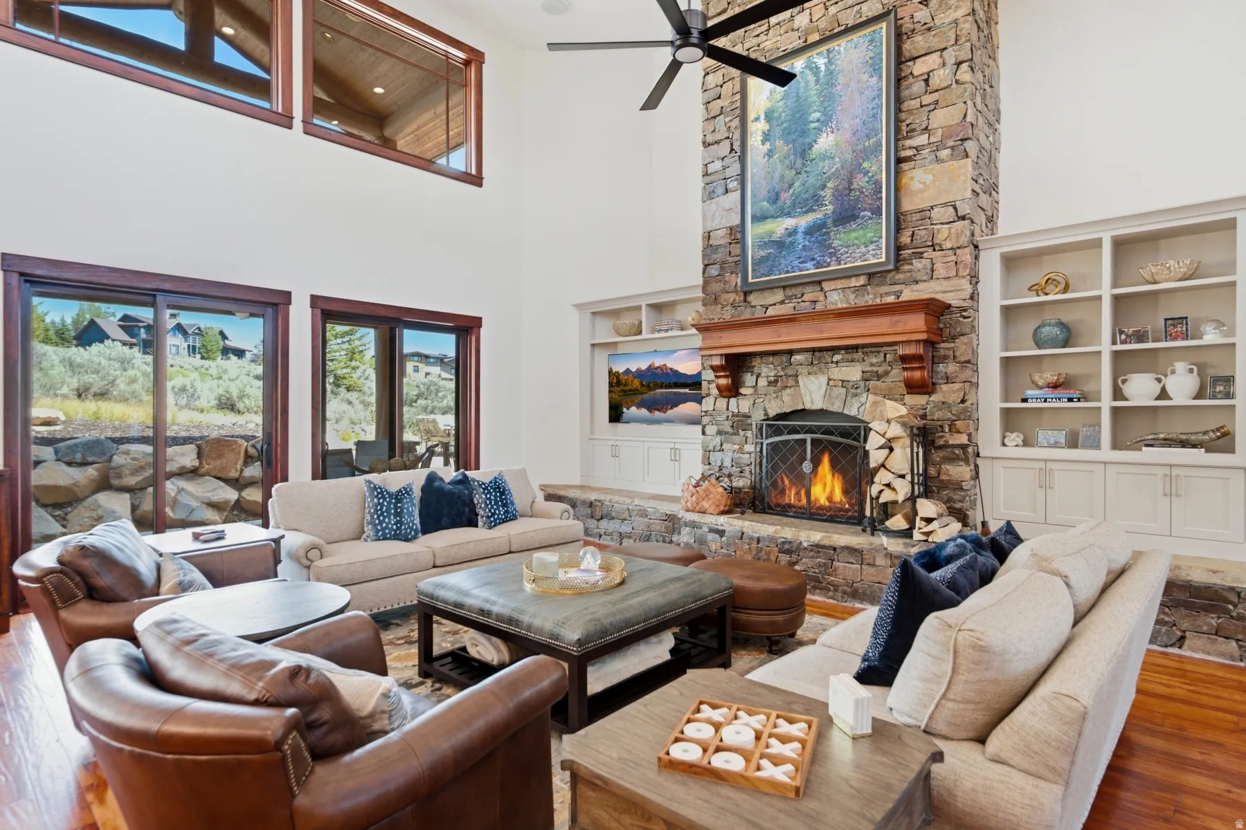 Spacious living area featuring a floor-to-ceiling stone fireplace, built-in shelving, vaulted ceilings, large windows, and hardwood flooring