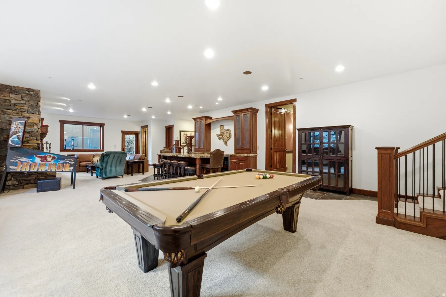 Spacious recreation room featuring a pool table, stone-clad fireplace, built-in wet bar with cabinetry, recessed lighting, and a wood staircase with wrought iron balusters