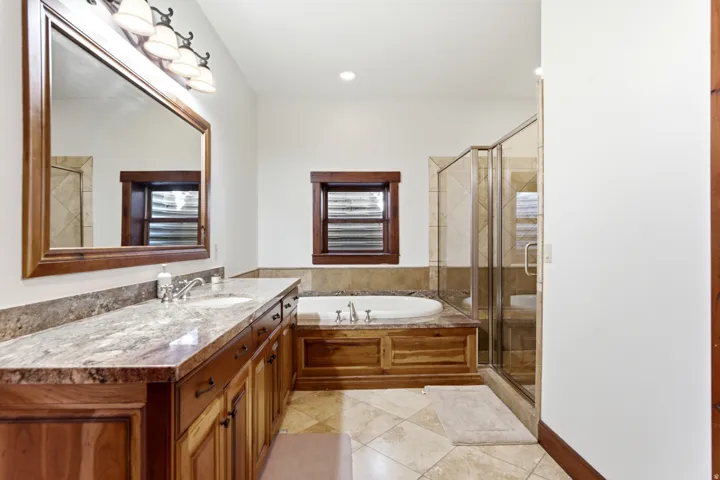 Spacious bathroom featuring a dual vanity with a stone countertop, a built-in jetted tub with wood paneling, a glass-enclosed shower, and decorative tile flooring