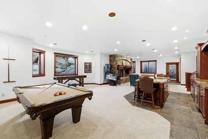 Expansive entertainment area featuring a billiard table, stone-clad fireplace, integrated bar with stone countertops, recessed lighting, and rich wood-trimmed windows