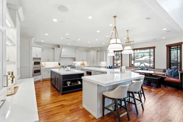 Spacious kitchen featuring two islands with white countertops, stainless steel double ovens, a built-in banquette, and warm wood-finish flooring