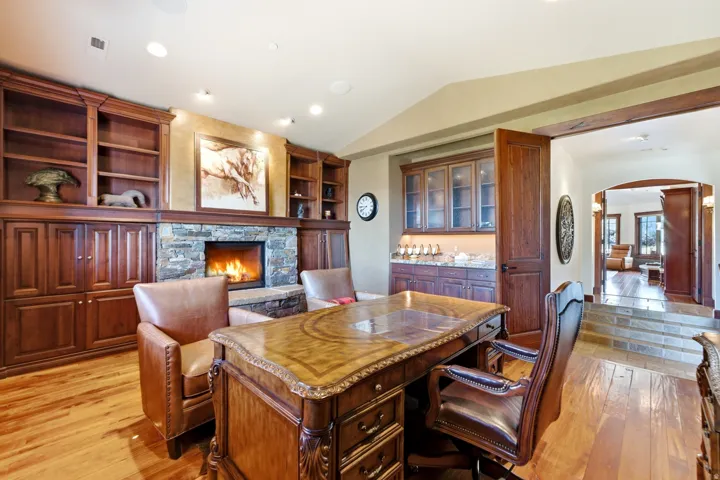 Home office featuring extensive wood built-in shelving and cabinetry, a stone fireplace, wood-finish flooring, and an integrated wet bar with glass-front upper cabinets