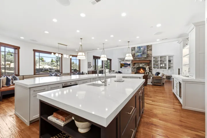 Spacious open-concept living area featuring two large kitchen islands with white countertops, warm wood-finish flooring, a stone fireplace, built-in shelving, and expansive windows