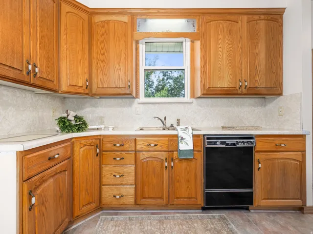 Kitchen featuring wood cabinetry, light countertops, patterned backsplash, stainless steel sink, and an integrated black dishwasher