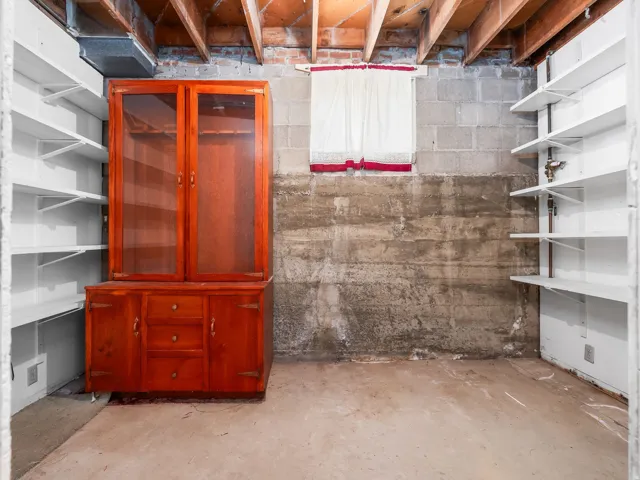 Storage area featuring exposed ceiling joists, concrete block walls, a small window opening, a built-in wood-finish cabinet with glass doors, and white shelving units