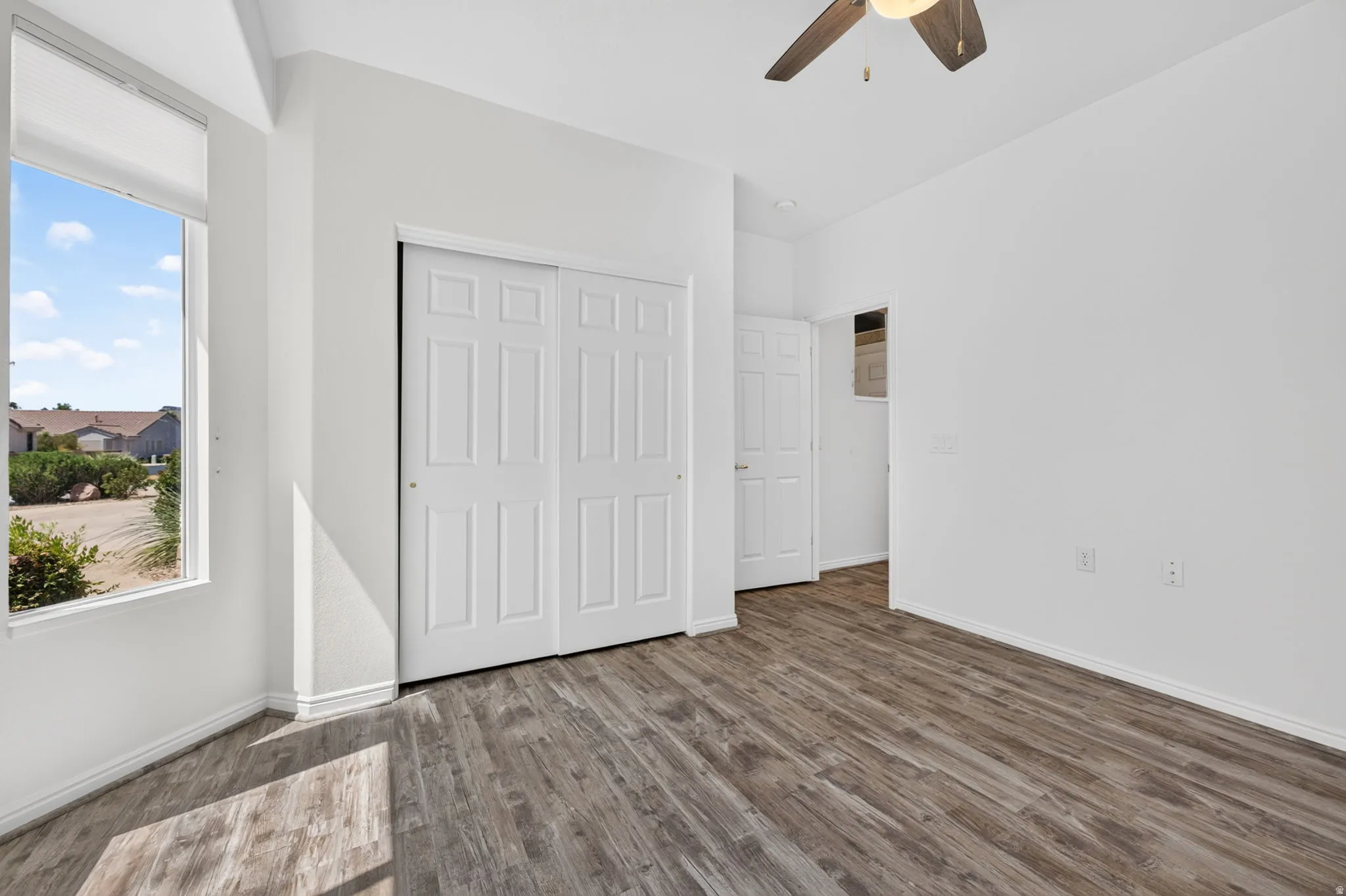 Room with wood-finish flooring, a ceiling fan with integrated lighting, and a window featuring a roller shade