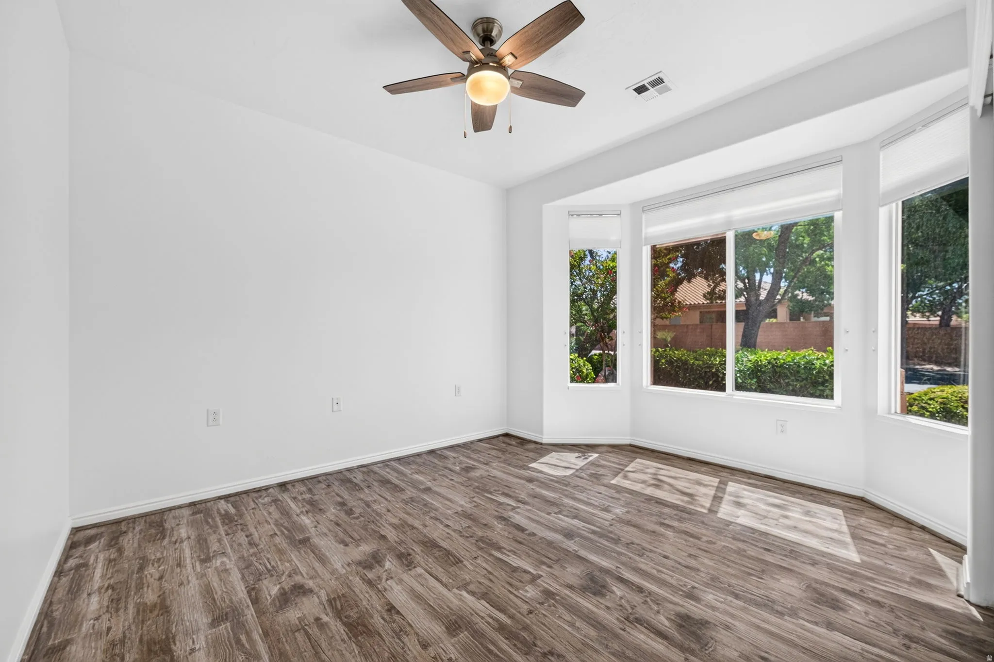 Spacious room featuring wood-finish flooring, a contemporary ceiling fan, and a multi-pane bay window with roller shades