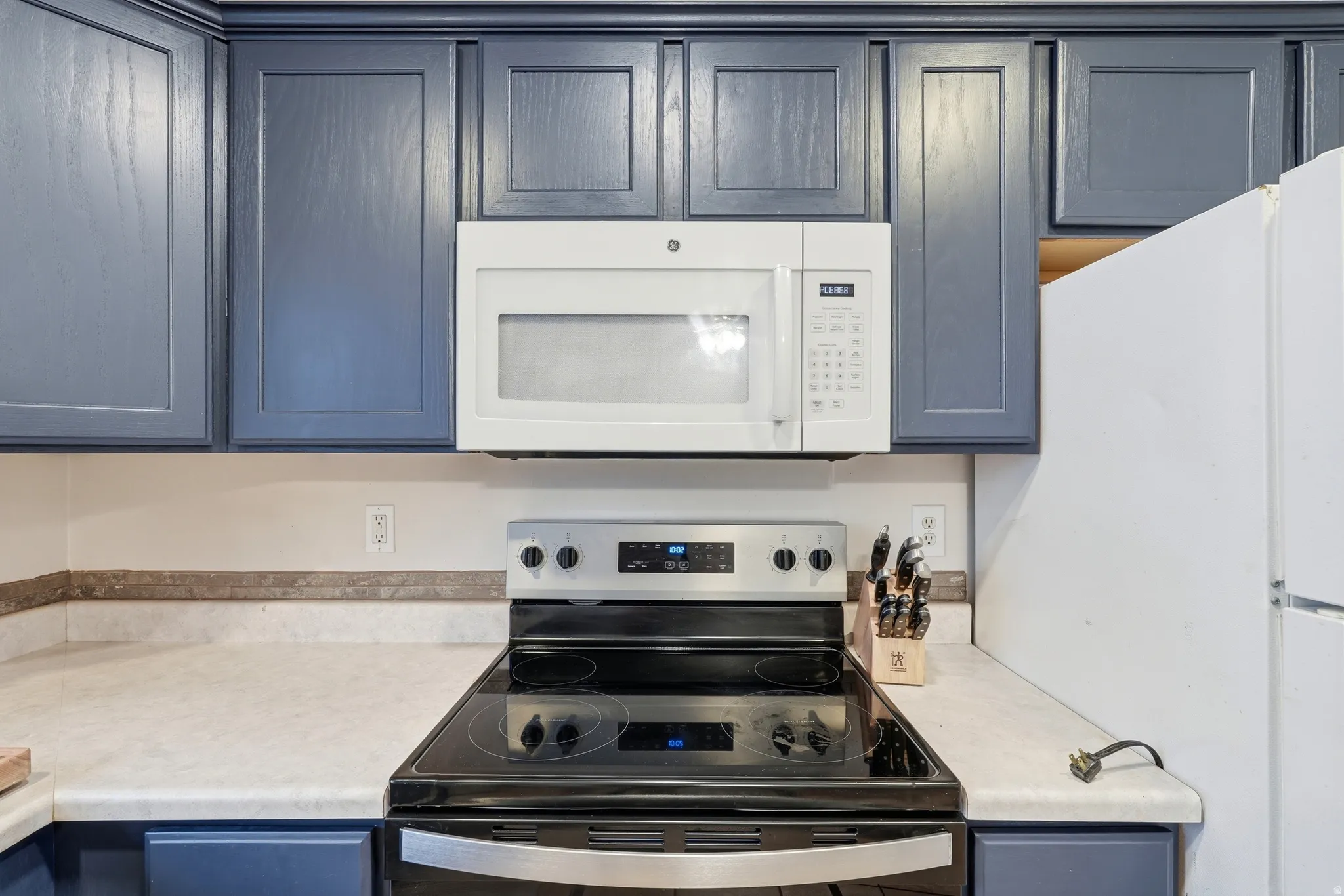 Kitchen featuring blue shaker-style cabinetry, white over-the-range microwave, electric range with stainless steel accents, light-toned countertops, and a partial view of a white refrigerator