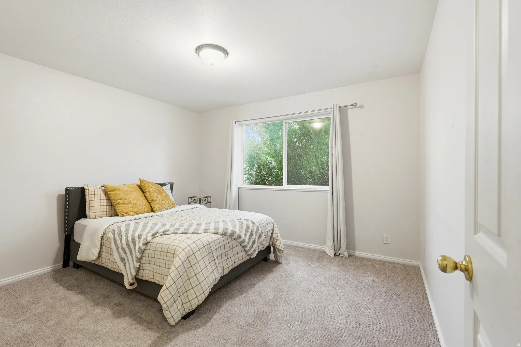 Spacious room featuring light-toned walls, a large window with exterior foliage visible, and carpet flooring
