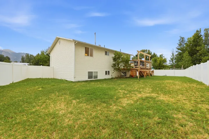 Expansive backyard featuring a white vinyl fence, a two-story residence with light-colored siding, a multi-level wood deck, and mature trees in the background