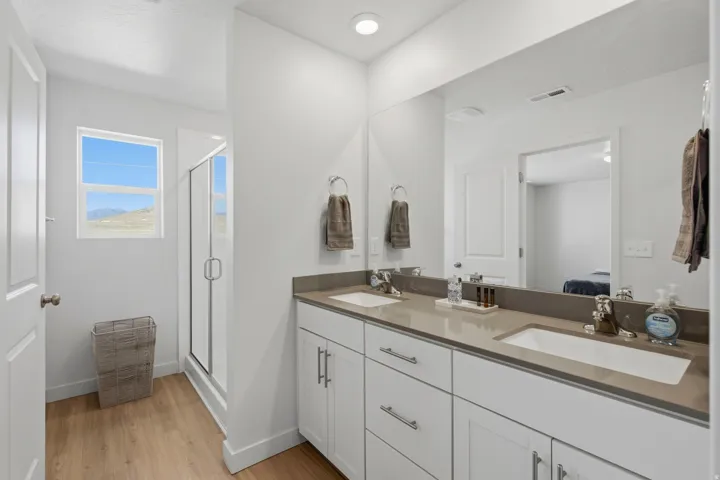 Bathroom featuring a dual vanity with undermount sinks, a large mirror, a glass-enclosed shower, and wood-finish flooring