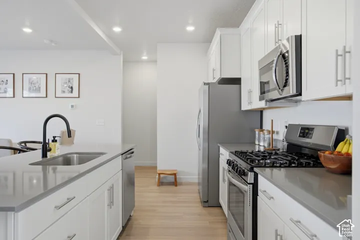 Bright kitchen featuring white cabinetry, stainless steel appliances, a center island with sink and matte black faucet, and wood-finish flooring