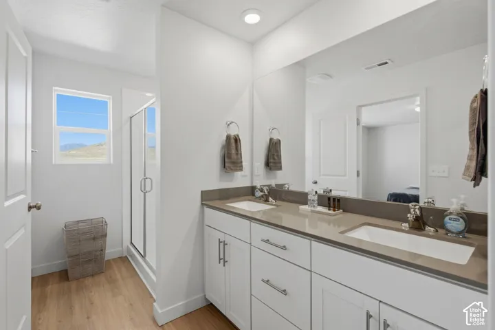 Bathroom featuring a dual vanity with undermount sinks, a large mirror, a glass-enclosed shower, and wood-finish flooring
