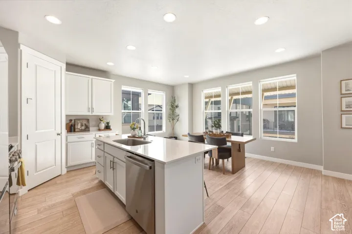 Kitchen and dining area featuring a central island with an undermount sink, stainless steel dishwasher, white cabinetry, light countertops, and wood-finish flooring