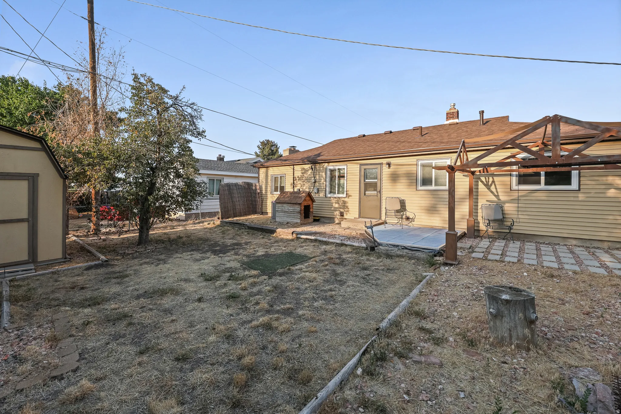 Spacious backyard featuring mature landscaping, gravel and paved hardscaping, pergola structure, and single-story home with siding exterior and attached garage
