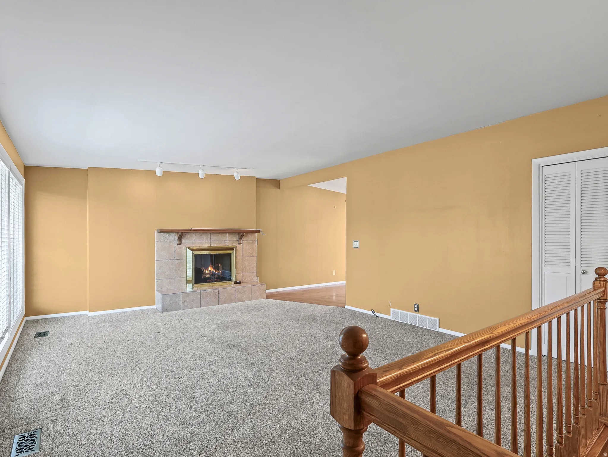 Carpeted living area featuring a tile-surround fireplace with a wood mantle, track lighting, and window blinds