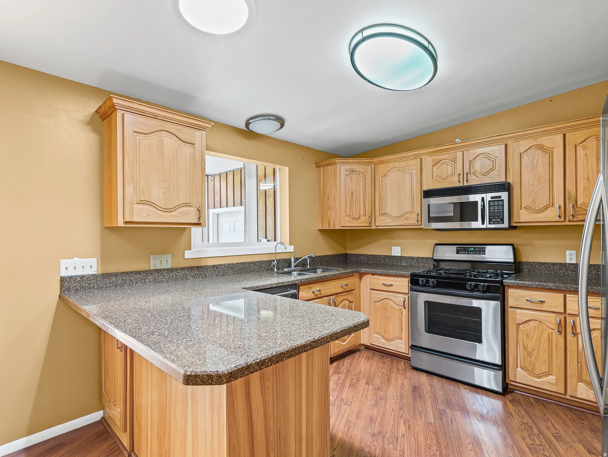 Kitchen featuring wood-finish flooring, light wood cabinetry, stone-look countertops, and stainless steel appliances including a gas range and microwave