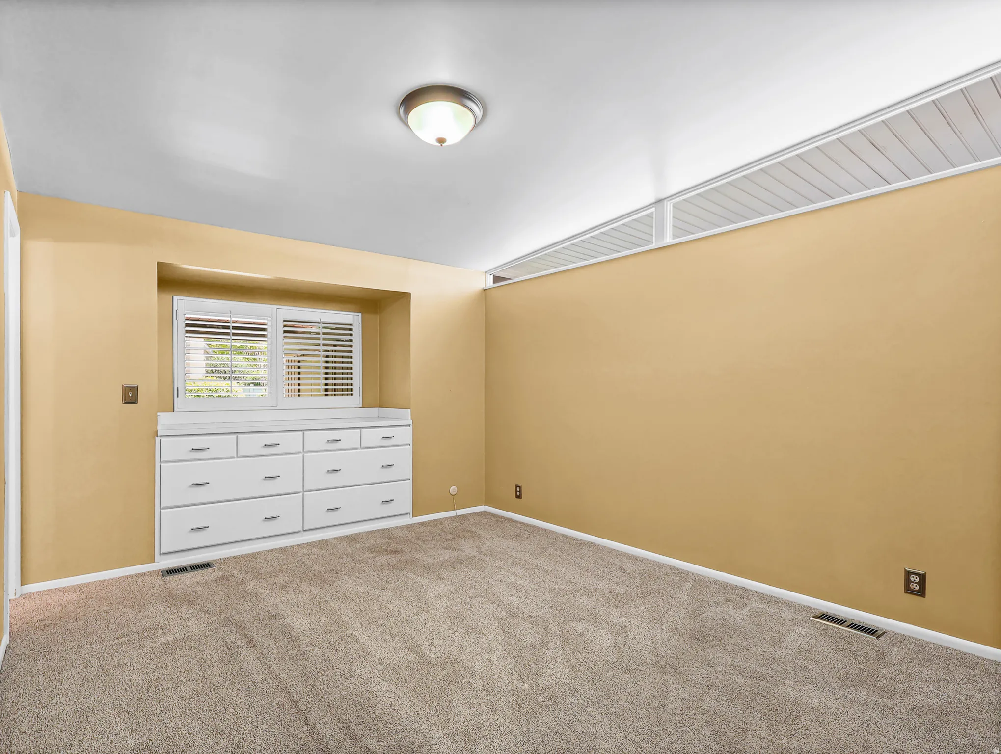 Carpeted room featuring a built-in dresser with nine drawers, a window with white louvered shutters, and a ceiling light fixture