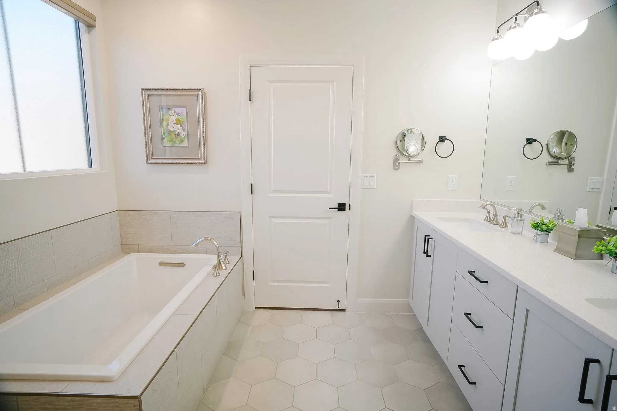 Bathroom featuring a built-in soaking tub with tile surround, a dual vanity with white countertops, a large wall mirror, and hexagonal floor tiling