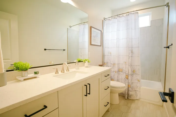 Bathroom featuring a dual-sink vanity with white cabinetry and black hardware, a light-toned countertop, a large wall-mounted mirror, a tiled shower-bath combination, and hexagonal floor tiling