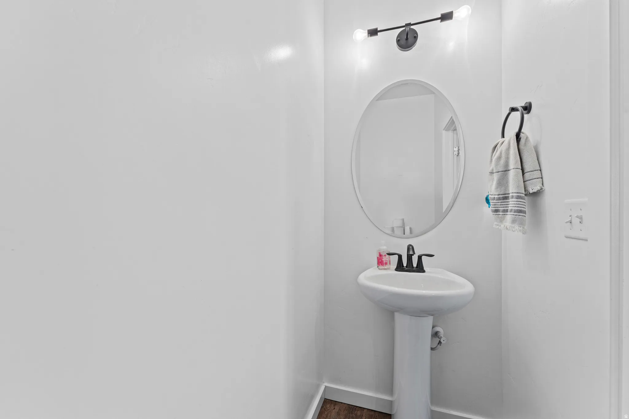 White pedestal sink with a matte black faucet, complemented by an oval mirror and a two-bulb vanity light fixture