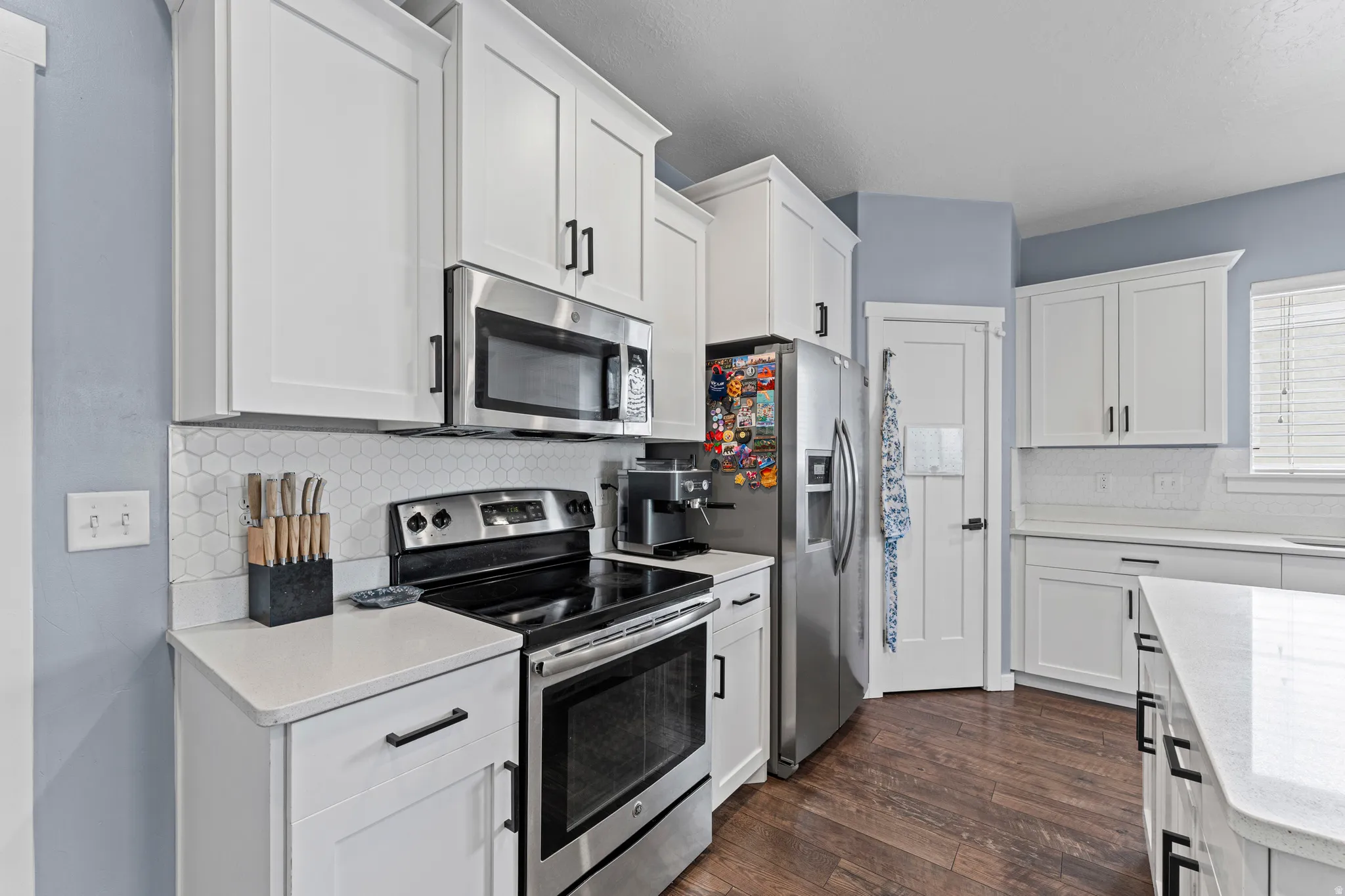 Contemporary kitchen featuring white shaker cabinetry, stainless steel appliances, white quartz countertops, hexagon tile backsplash, and wood-finish flooring