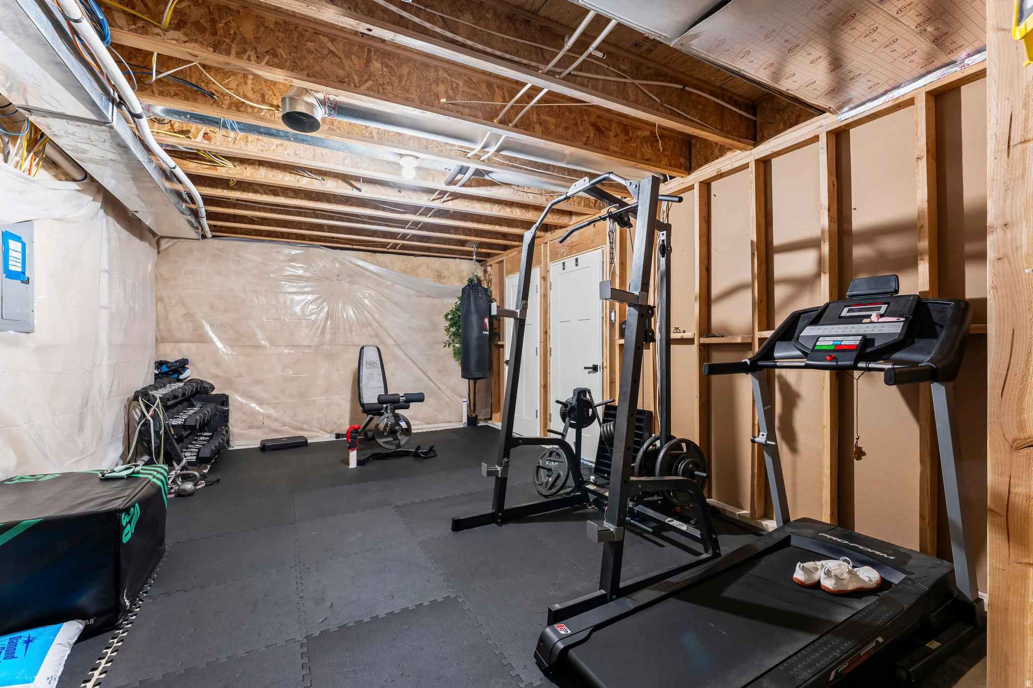 Unfinished basement with exposed wood framing and ceiling joists, featuring resilient black flooring tiles, a white paneled door, and exposed electrical conduit