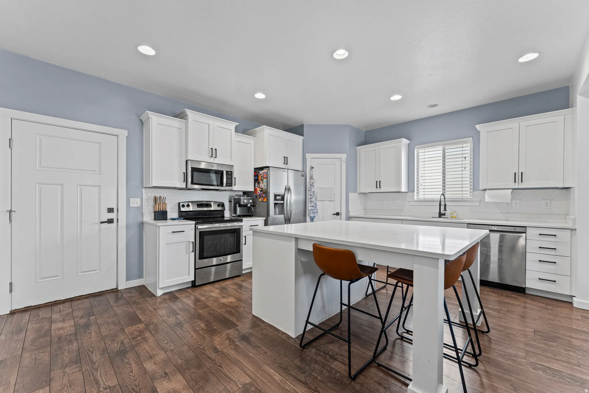 Modern kitchen featuring white shaker cabinetry, stainless steel appliances, a large center island with a solid surface countertop, wood-finish flooring, and recessed lighting