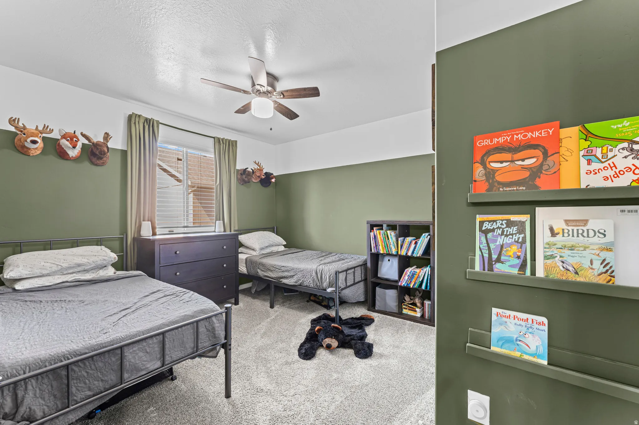 Carpeted room featuring a ceiling fan with integrated lighting, a window with blinds and drapes, and a built-in shelving unit