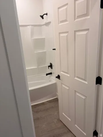 Bathroom featuring a white tub/shower combination with integrated shelving, matte black fixtures, and wood-finish flooring
