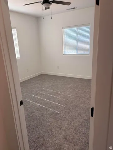 Spacious room featuring gray carpet flooring, two windows with horizontal blinds, a ceiling fan with integrated lighting, and white baseboards