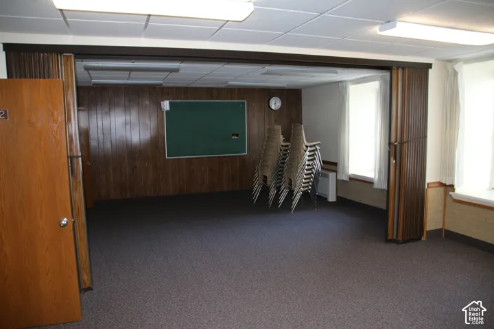 Spacious room featuring wood-finish wall paneling, a drop ceiling with fluorescent lighting, and two windows with white curtains