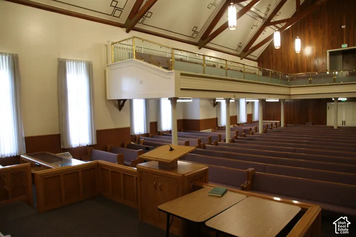 Spacious hall featuring vaulted wood-beam ceilings, an upper-level balcony with a brass railing, and multiple window openings with sheer curtains