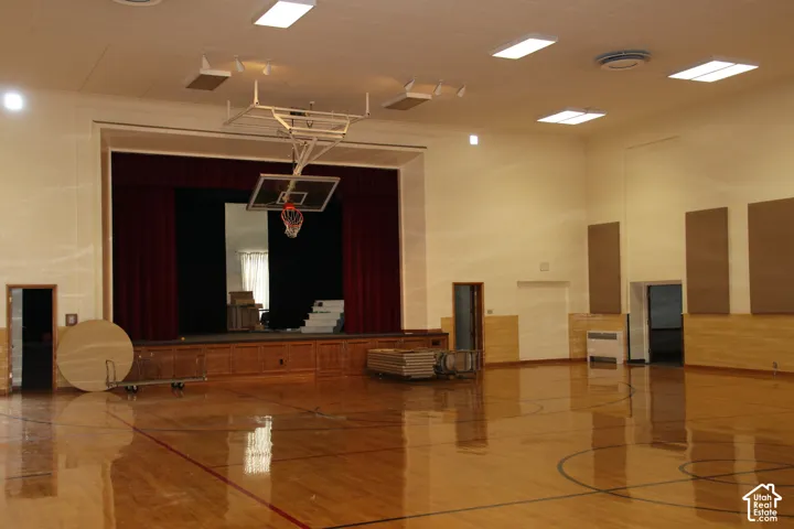 Spacious multi-purpose room featuring a hardwood floor with court markings, a permanent stage with red curtains, and an adjustable basketball hoop