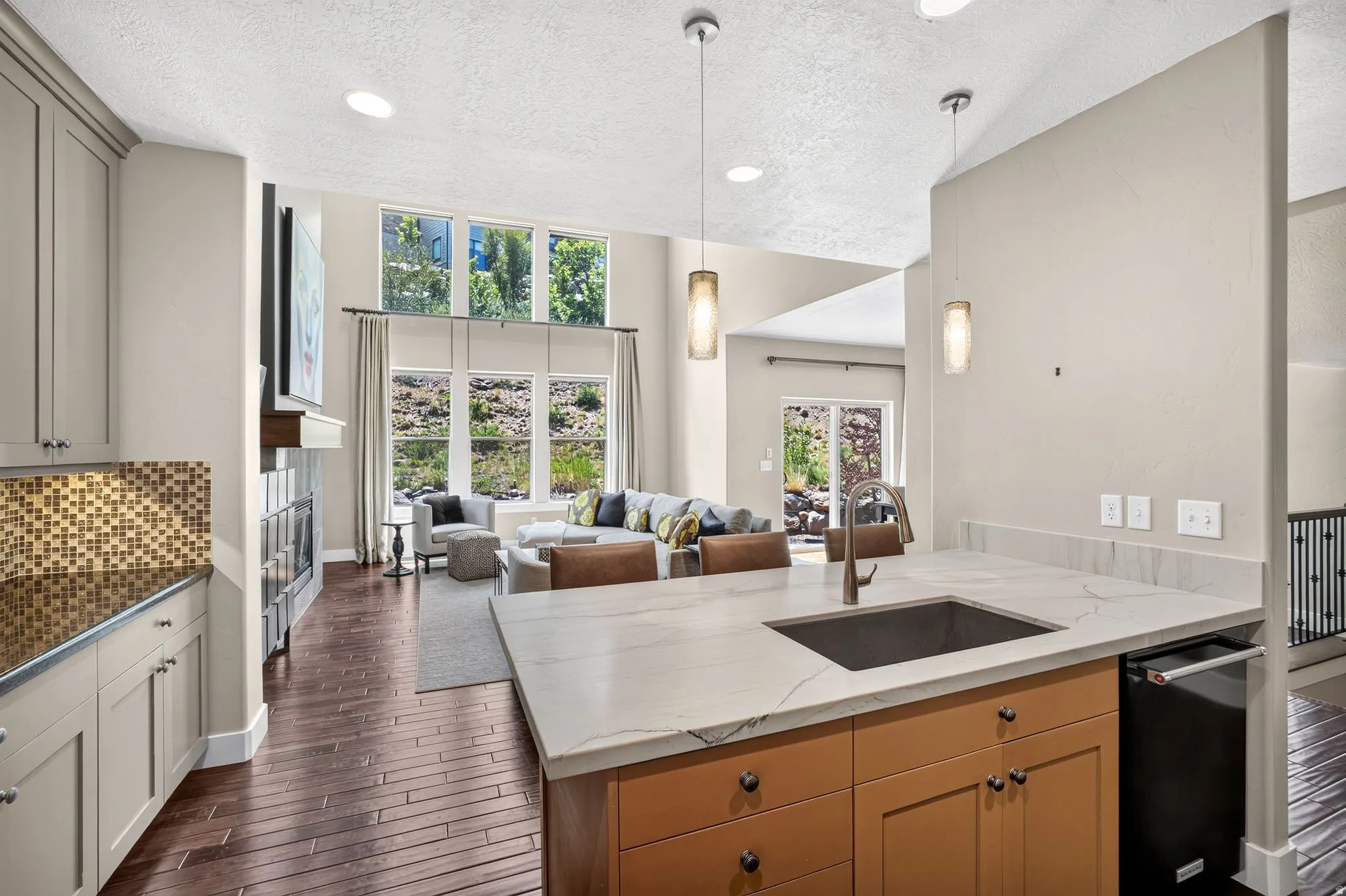 Open-concept living area featuring a kitchen island with an undermount sink and pendant lighting, wood-finish flooring, a contemporary fireplace, and large windows providing natural light